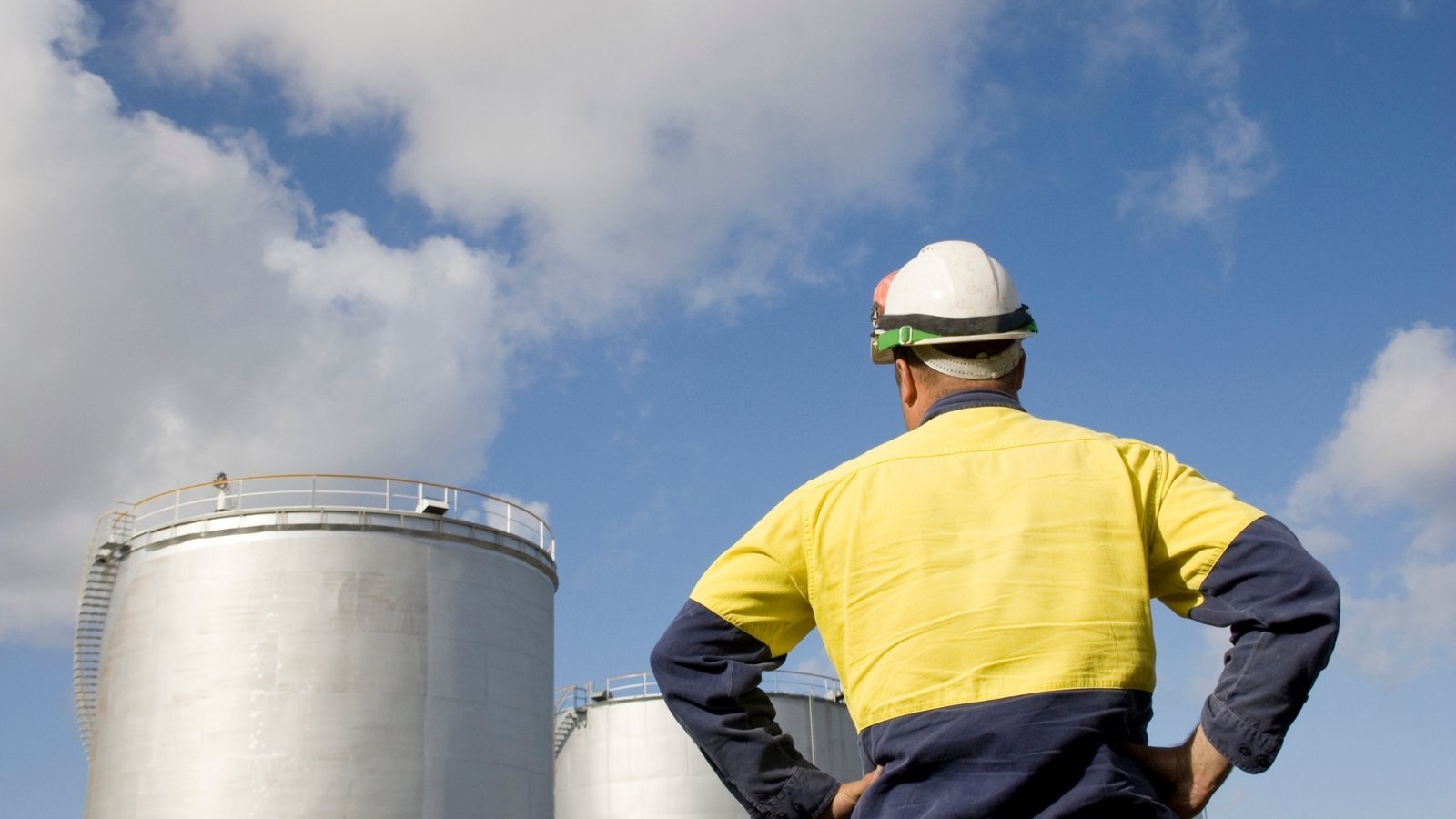 A worker in a hard hat looking up at large aboveground crude storage tanks