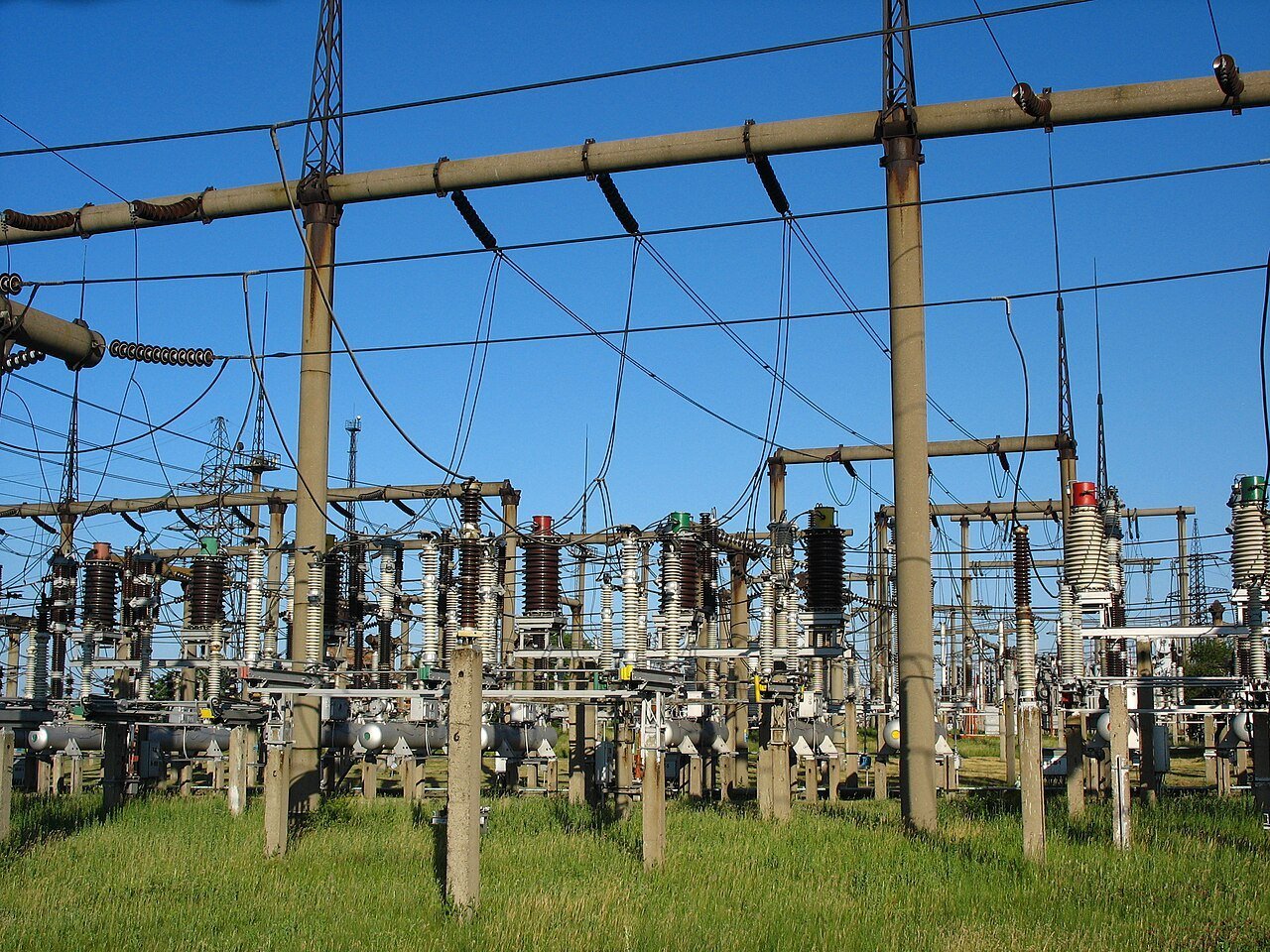 Outdoor high voltage electrical substation switchyard, with steel gantries, busbars, disconnect switches and porcelain insulators standing in open grass under a clear blue sky