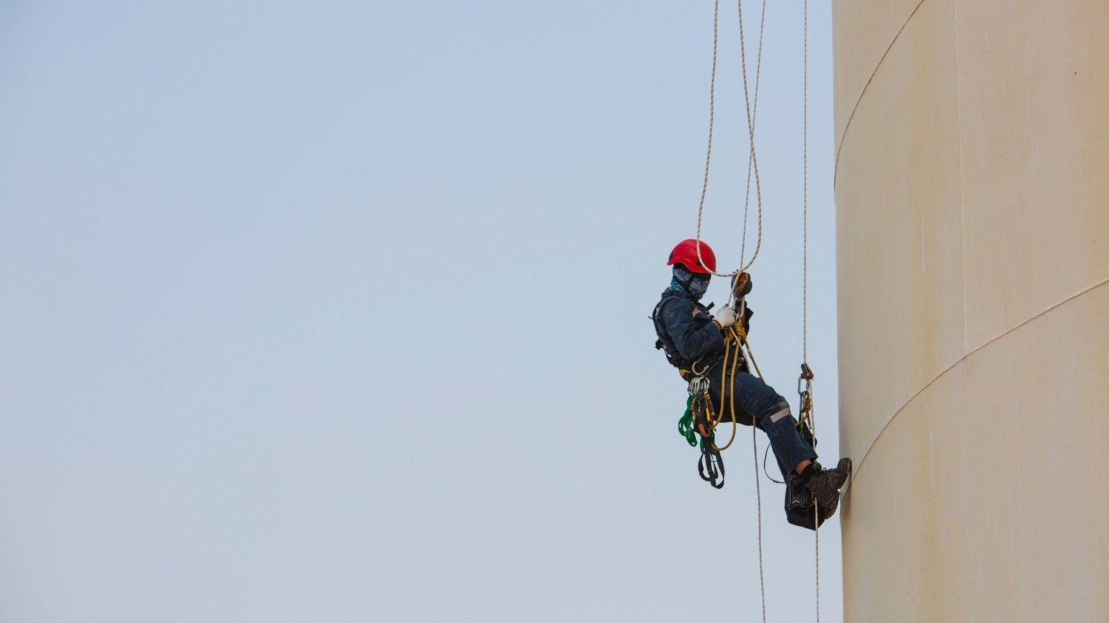 A technician in a red helmet abseiling down the curved white wall of a large storage tank