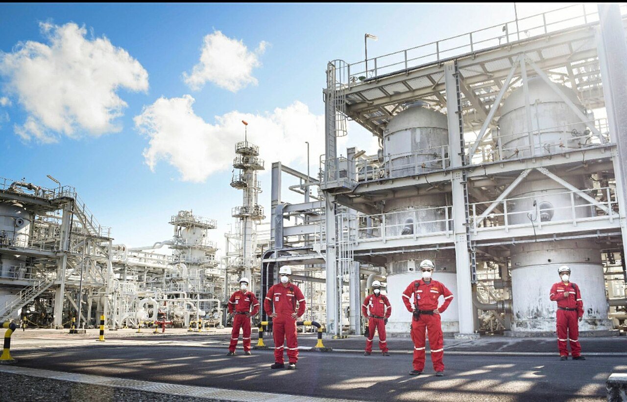 Six operators in red coveralls and white hard hats standing in front of a large steel process vessel at an onshore gas processing plant