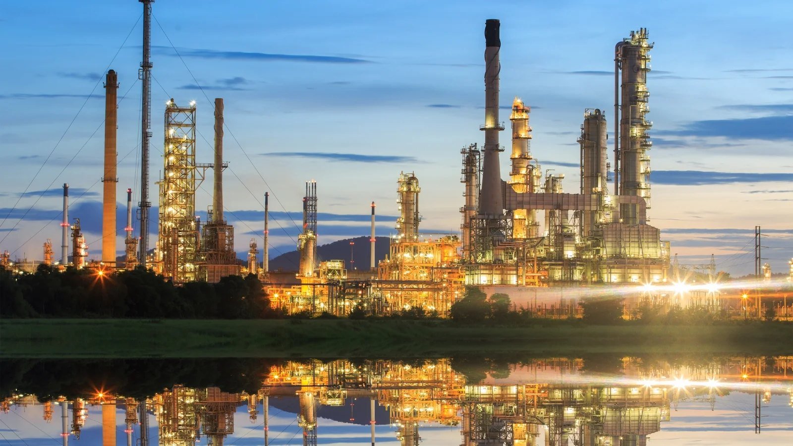 A large refinery and petrochemical complex lit up at dusk, its stacks, columns and flare towers reflected in still water in the foreground