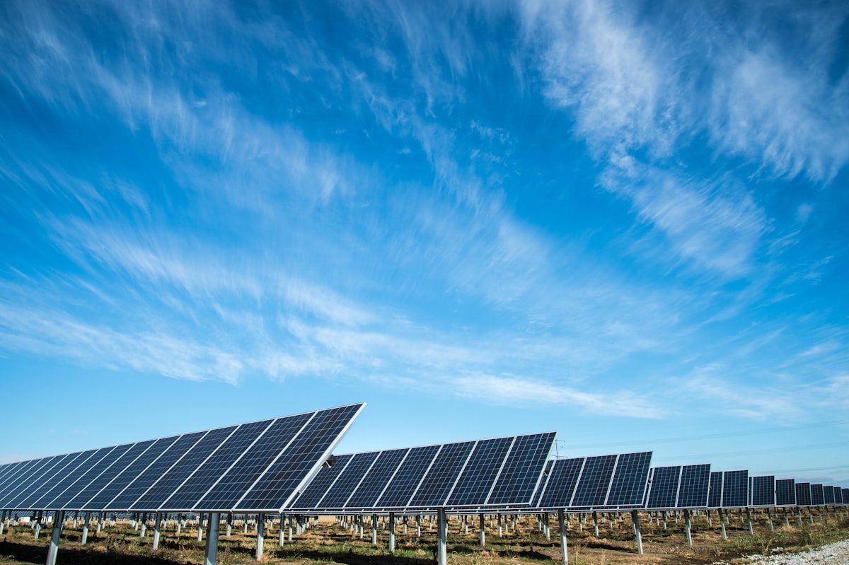 A long row of utility scale solar tracker panels receding to the horizon in open country under a wide blue sky